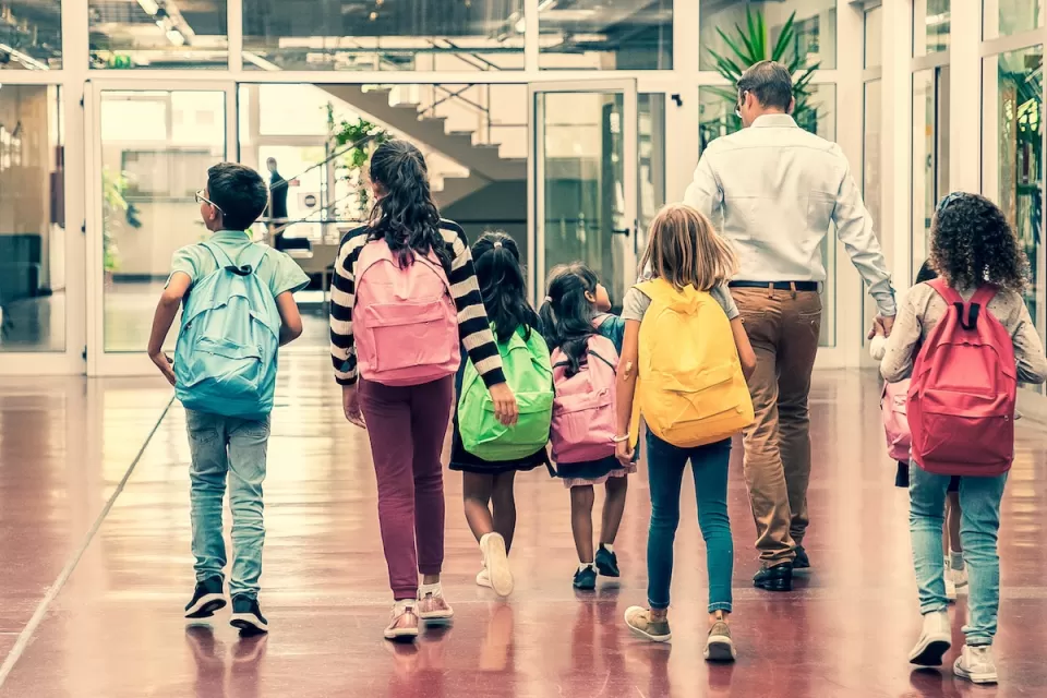 Back view of group of six children and one adult walking down school hallway.
